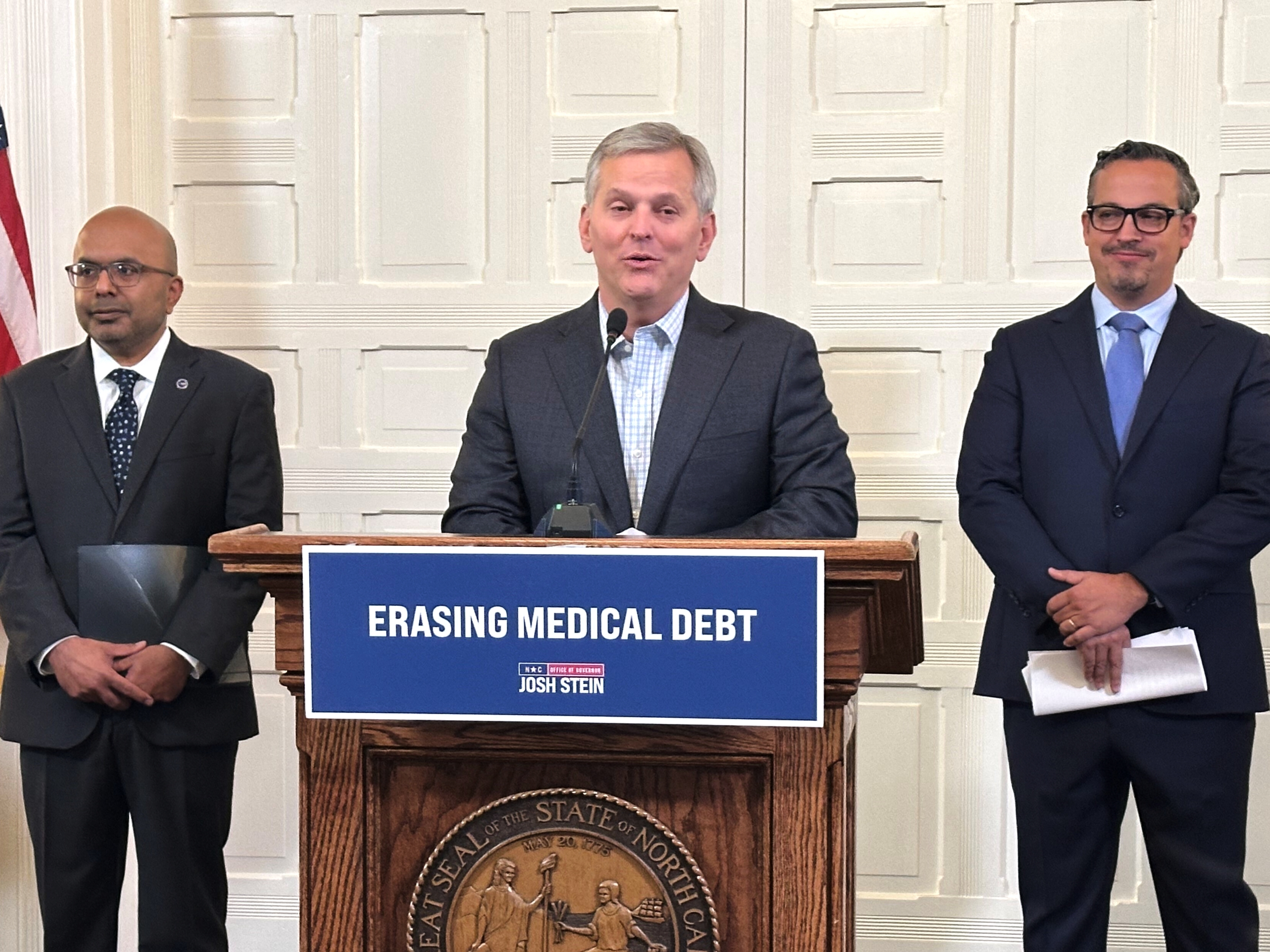 caption: North Carolina Gov. Josh Stein (center), flanked by the state's health secretary, Dr. Dev Sangvai (left) and an executive from Undue Medical Debt, Jose Penabad, speaks about the elimination of medical debt through an initiative involving hospitals and Medicaid in Raleigh, N.C.