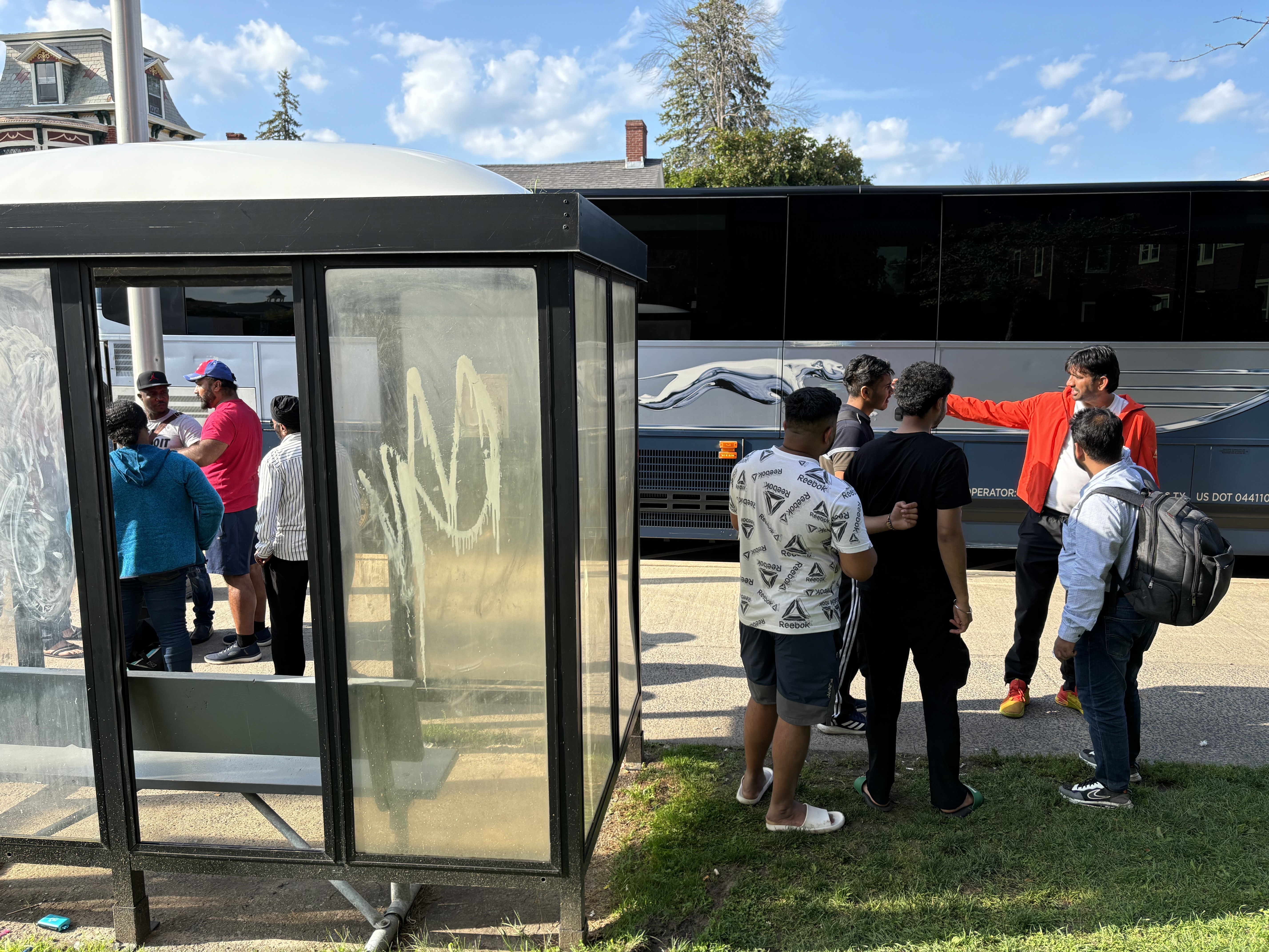 caption: A group of Indian and Haitian immigrants arrive at a bus stop in Plattsburgh, N.Y. on a Saturday afternoon in August. The migrants were received by Indian drivers who take them to New York City for a fee.