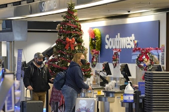 caption: Travelers wear masks as they check in next to a holiday tree at a Southwest Airlines ticket desk, Friday, Dec. 10, 2021, at Seattle-Tacoma International Airport in Seattle. 