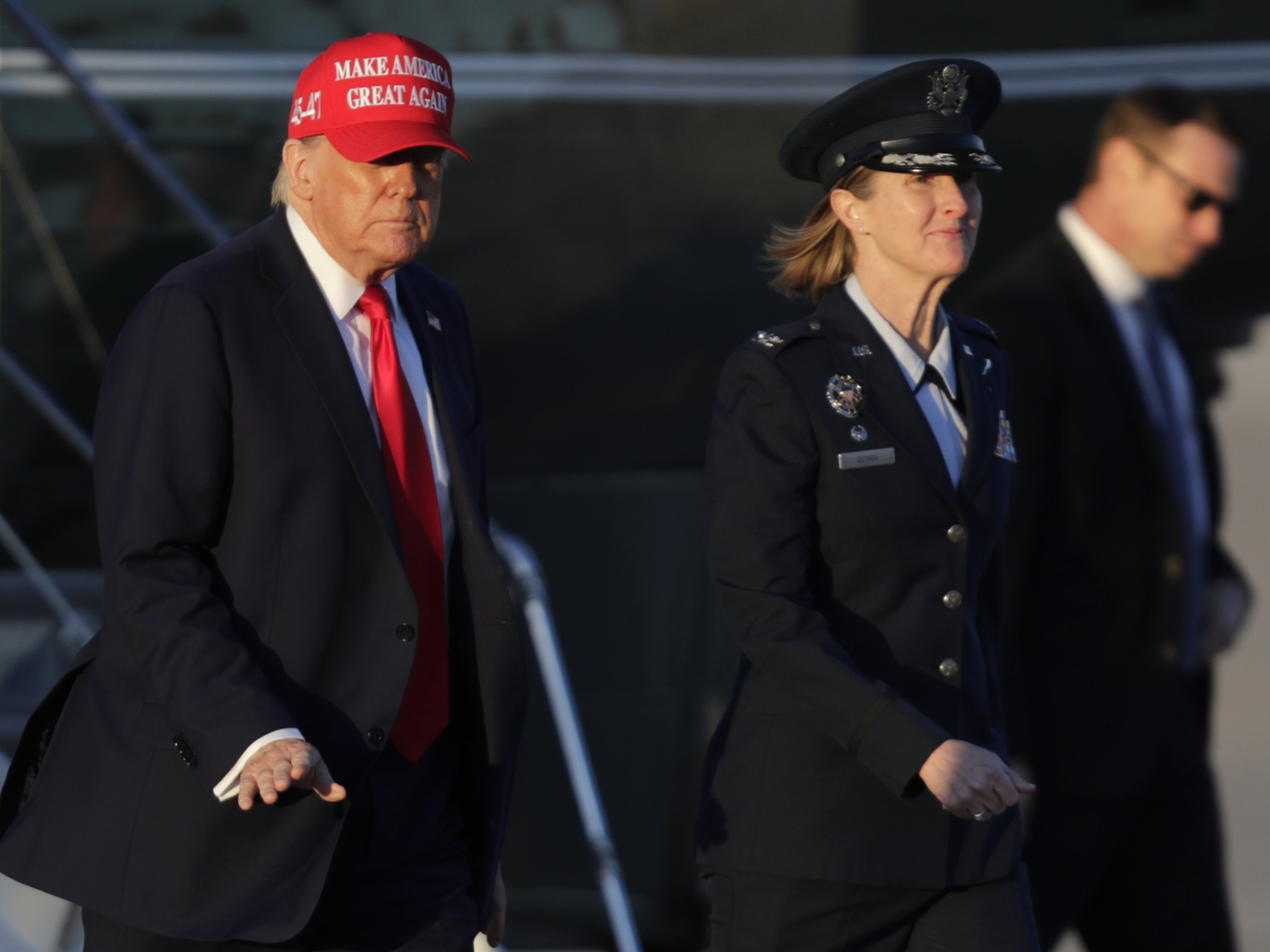 caption: President Donald Trump, left, gestures as is escorted by Air Force Col. Angela Ochoa, commander of the 89th Airlift Wing, center, as he walks from Marine One before boarding Air Force One, Friday, Feb. 28, 2025, at Joint Base Andrews, Md.
