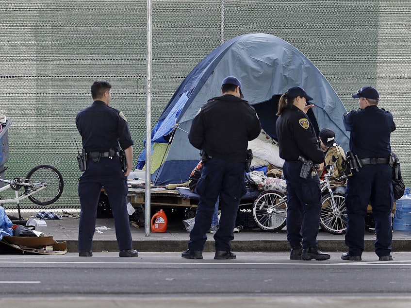 caption: San Francisco police officers wait while homeless people collect their belongings. Nearly a quarter of the country's homeless population lives in California.