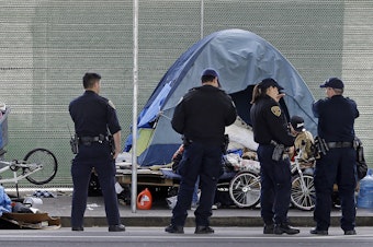 caption: San Francisco police officers wait while homeless people collect their belongings. Nearly a quarter of the country's homeless population lives in California.