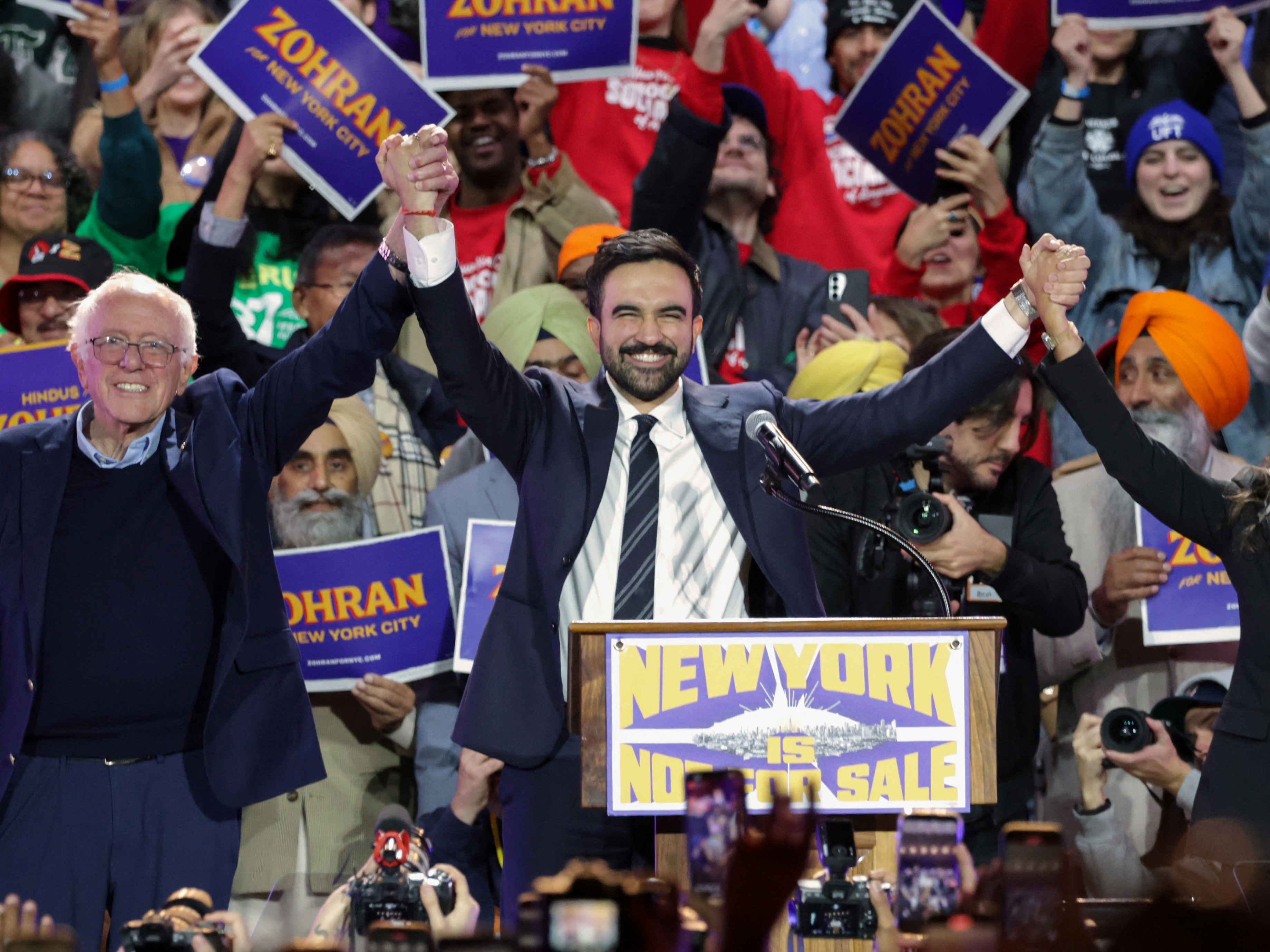 caption: New York City Mayor-elect Zohran Mamdani holds hands with Sen. Bernie Sanders and Rep. Alexandria Ocasio-Cortez at a campaign rally in October. All three identify as democratic socialists.