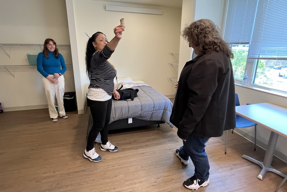 caption: Plymouth Housing case manager Angel Cezear smudges the room with burning sage, at the request of new resident Kevin Thomas Kiso (right) as his Salvation Army caseworker, Katy Cossette (left) looks on.