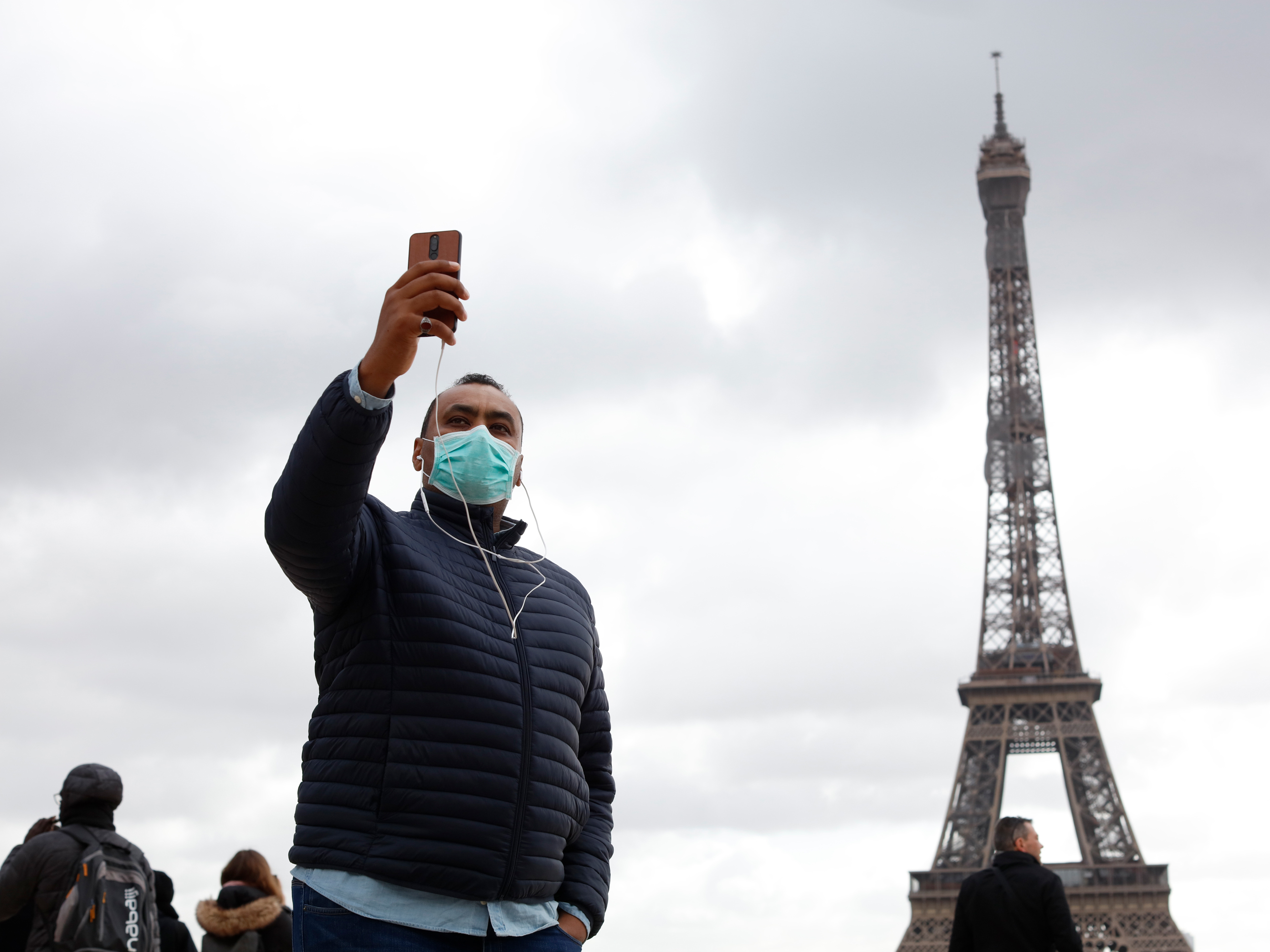 caption: People with protective masks walk in front of the Eiffel Tower in Paris on March 9. The new coronavirus has had an impact on international travel.