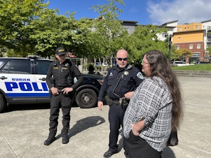 caption: Burien Police Officer Mark Hayden, center, asks Cydney Moore, right, to inform homeless campers about trash pickups and upcoming street fairs in downtown Burien. 