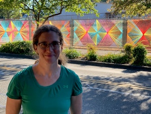 caption: Christy Caravaglio smiles proudly in front of her public art installation in Burien. Sunshine makes the already bright colors look almost luminescent. Christy weaves yarn into the chain link fences she passes daily in an effort to brighten up the perpetually gray landscape. 