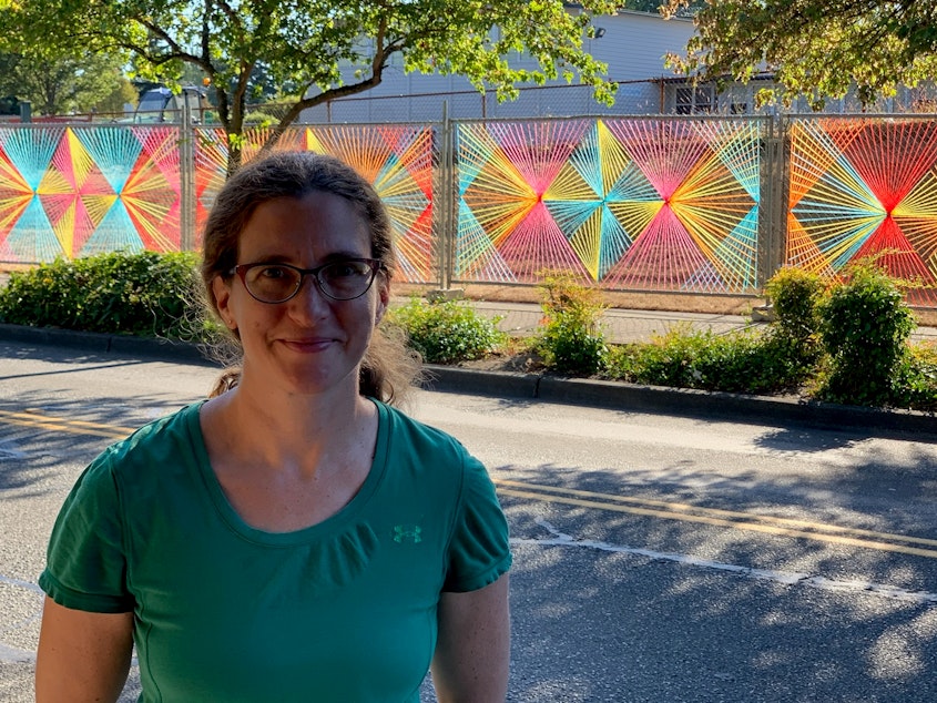 caption: Christy Caravaglio smiles proudly in front of her public art installation in Burien. Sunshine makes the already bright colors look almost luminescent. Christy weaves yarn into the chain link fences she passes daily in an effort to brighten up the perpetually gray landscape.
