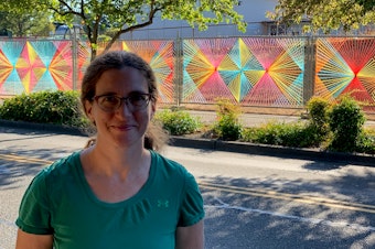 caption: Christy Caravaglio smiles proudly in front of her public art installation in Burien. Sunshine makes the already bright colors look almost luminescent. Christy weaves yarn into the chain link fences she passes daily in an effort to brighten up the perpetually gray landscape. 