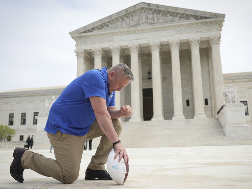 caption: Joseph Kennedy, seen here taking a knee in front of the U.S. Supreme Court last spring, will return to coach at Bremerton High School. His practice of praying on the field sparked a court case over accommodating religious expression in public schools.