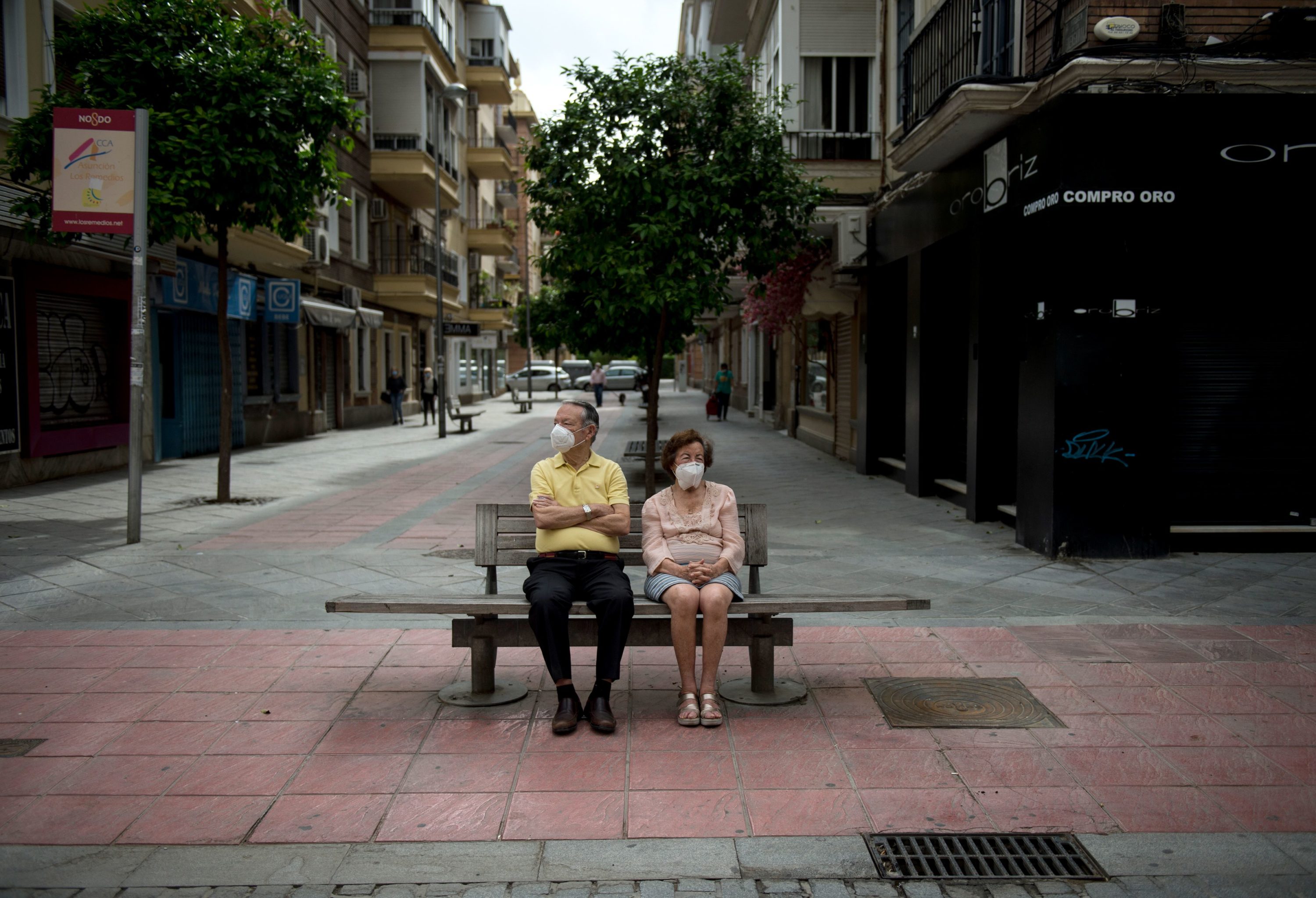 caption: An elderly couple wearing face masks sits on a bench in Seville on May 7, 2020. (CRISTINA QUICLER/AFP via Getty Images)