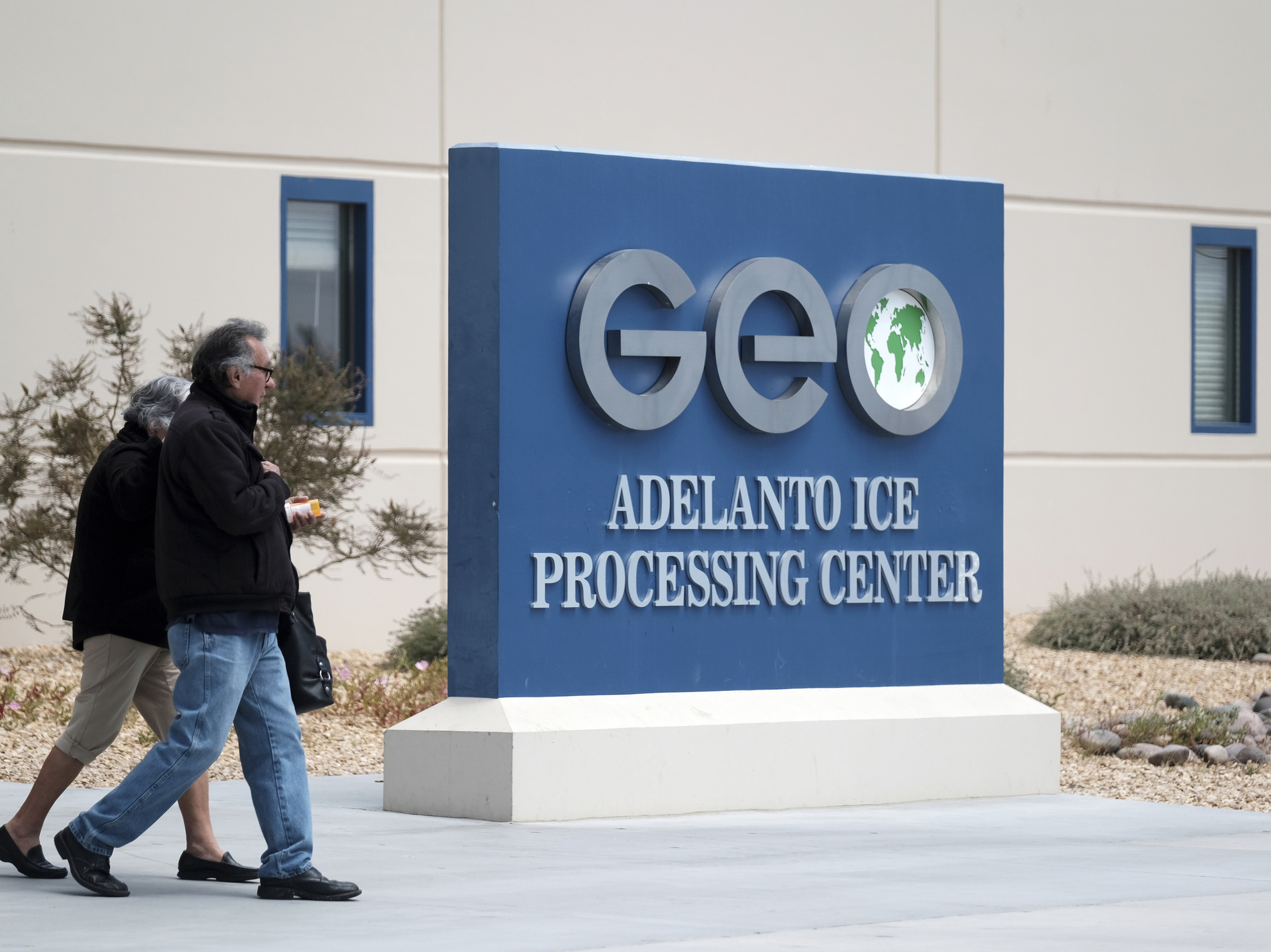 caption: People walk outside the U.S. Immigration and Enforcement Processing Center operated by GEO Group Inc. in Adelanto, Calif. The company is facing a lawsuit for the alleged use of hazardous chemicals in the facility during the COVID-19 pandemic.