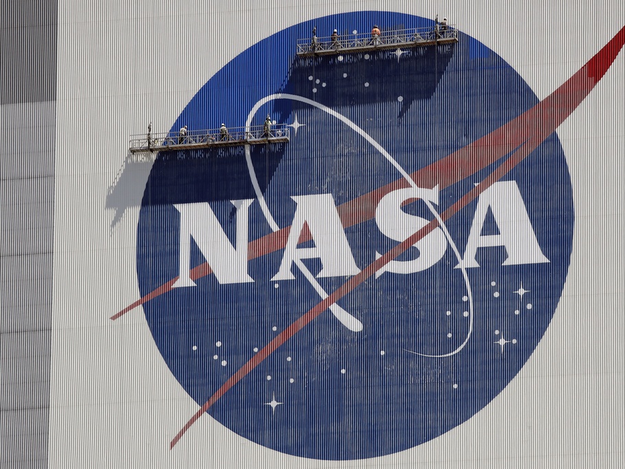 caption: Workers on scaffolding repaint the NASA logo near the top of the Vehicle Assembly Building in 2020 at the Kennedy Space Center in Cape Canaveral, Fla.