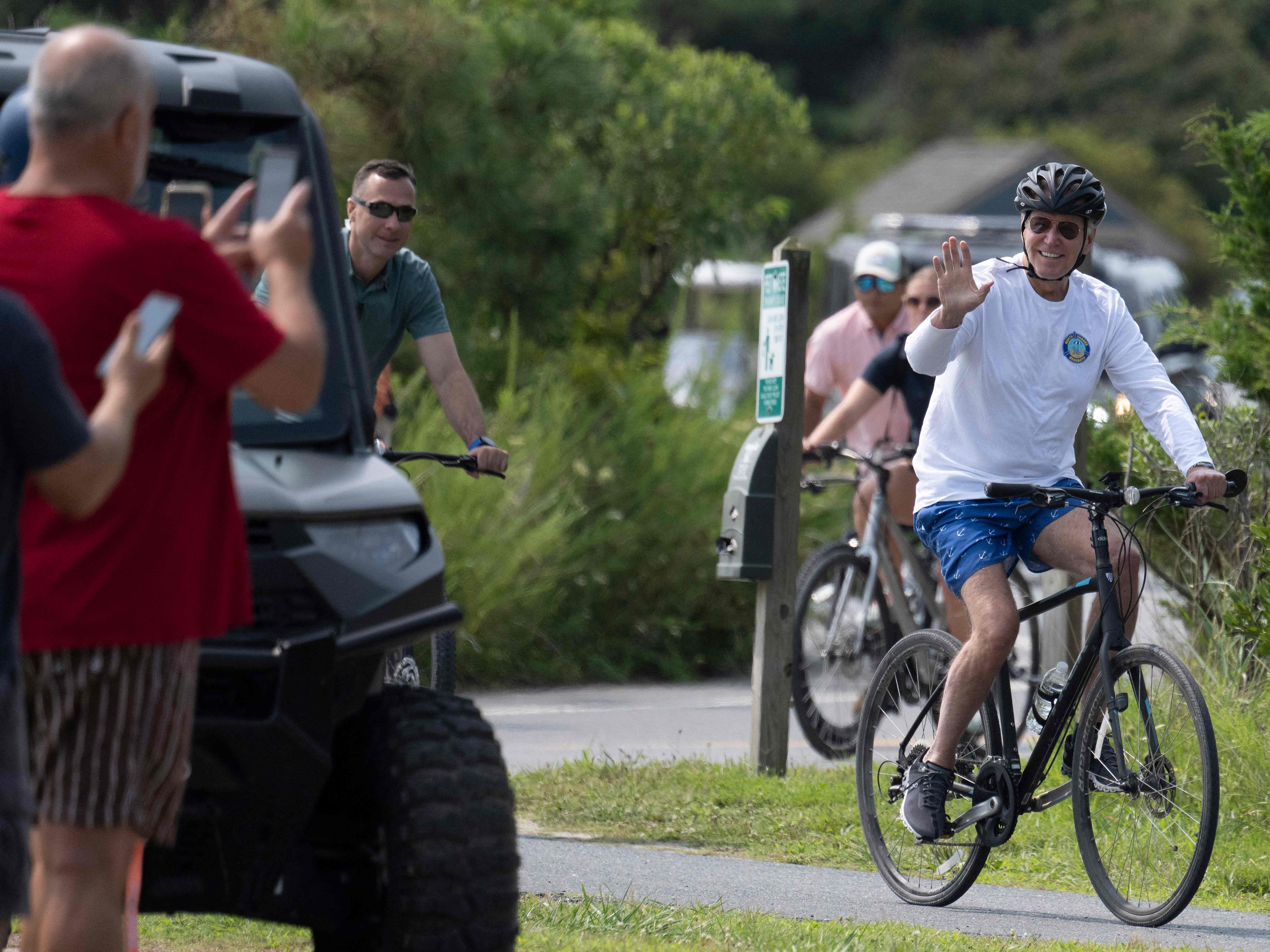 caption: President Biden waves to onlookers as he rides his bike through Gordons Pond State Park in Rehoboth Beach, Del., on Tuesday.