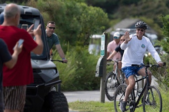 caption: President Biden waves to onlookers as he rides his bike through Gordons Pond State Park in Rehoboth Beach, Del., on Tuesday.