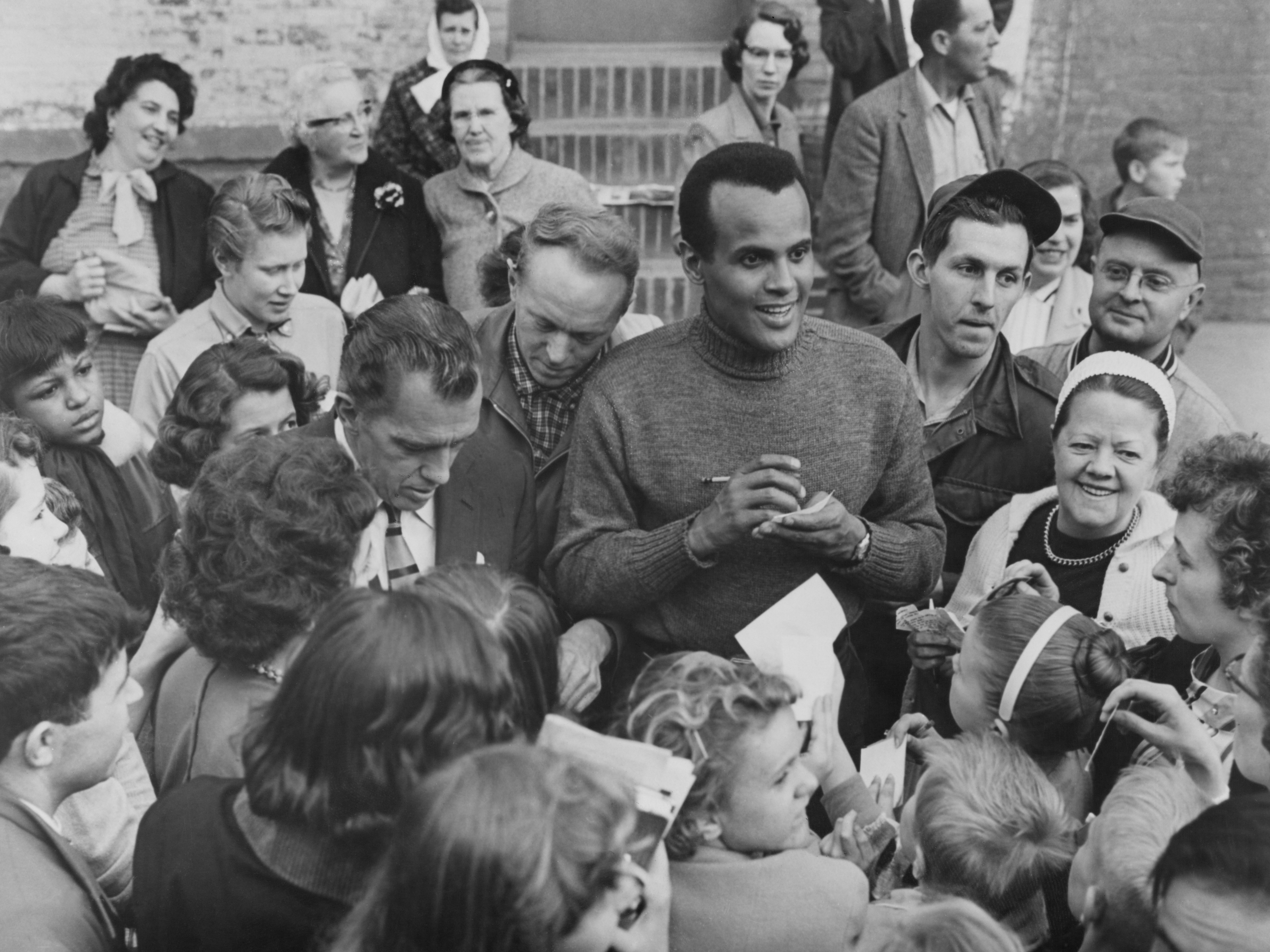 caption: Harry Belafonte, alongside Ed Sullivan, signs autographs for fans outside CBS Studio 50 in New York City, circa 1955.