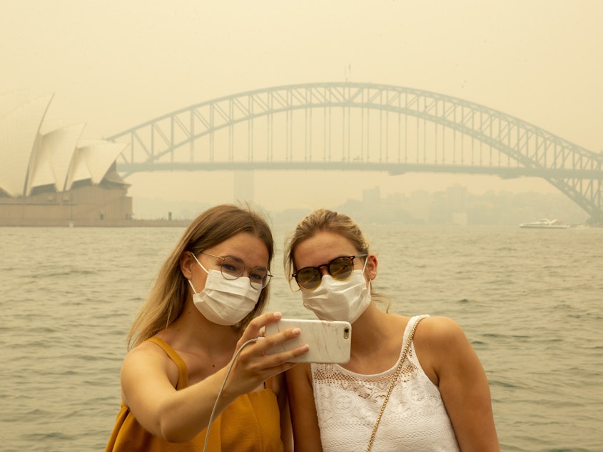 caption: Tourists, wearing face masks due to heavy smoke, take a selfie at Sydney Harbour on Dec. 19, 2019. As news of Australia's wildfires spreads around the world, fewer tourists are arriving in the country.