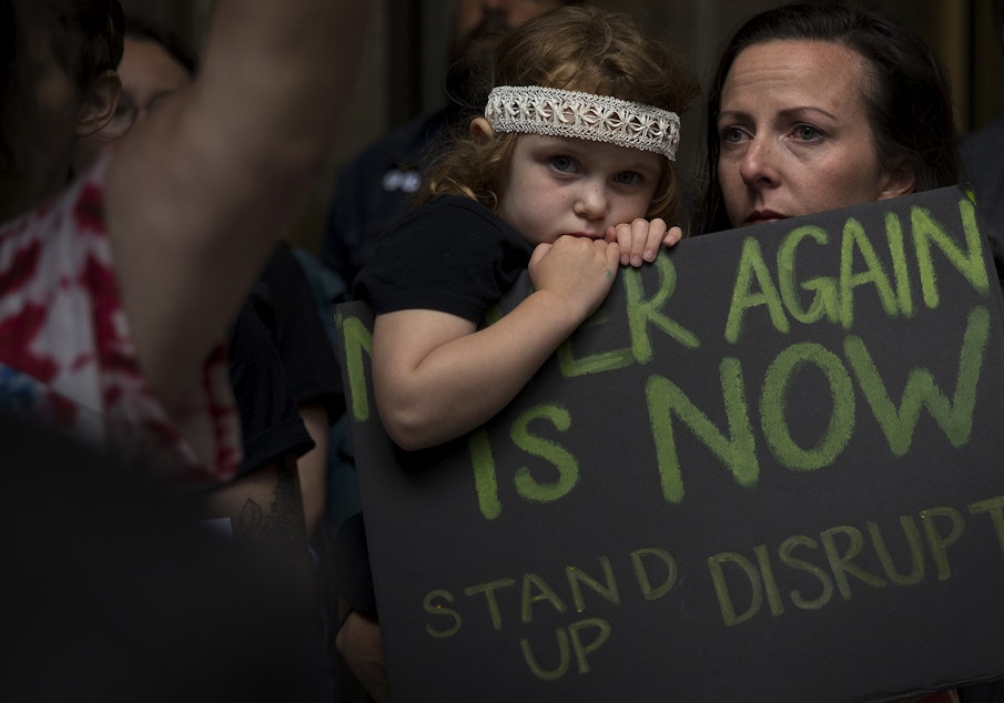 caption: Gloria Dykstra and Pearl Dykstra, 4, join hundreds of others in protest, holding a sign that reads 'Never Again is Now, Stand up and Disrupt,' outside of Seattle's downtown ICE offices on Thursday, August 8, 2019, on 2nd Avenue in Seattle.