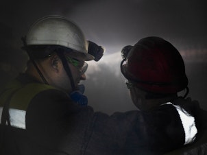caption: Mine workers are surrounded by dust as a drill bit chews into the wall of the mine.