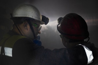 caption: Mine workers are surrounded by dust as a drill bit chews into the wall of the mine.