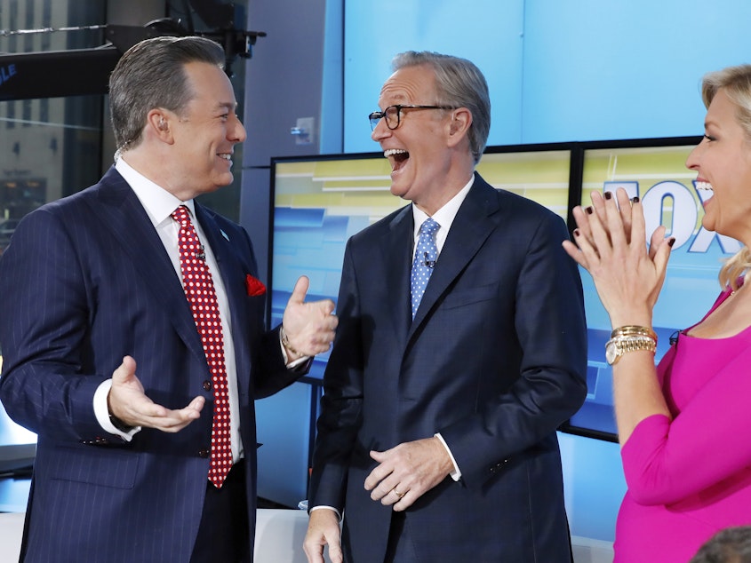 caption: Fox News has fired former anchor Ed Henry, left, who is standing with co-hosts Steve Doocy and Ainsley Earhardt on the "Fox & friends" television program in September 2019. (AP Photo/Richard Drew)