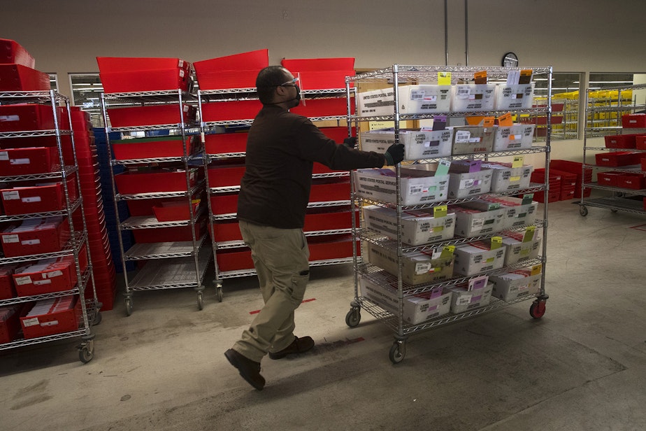 caption: An election worker moves a rack full of sorted ballots on Wednesday, October 28, 2020, at King County Elections in Renton.