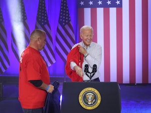 caption: President Biden puts on a United Auto Workers T-shirt in Belvidere, Ill., on Nov. 9, 2023. Biden on Tuesday is announcing new tariffs on imports of Chinese goods, including electric vehicles.