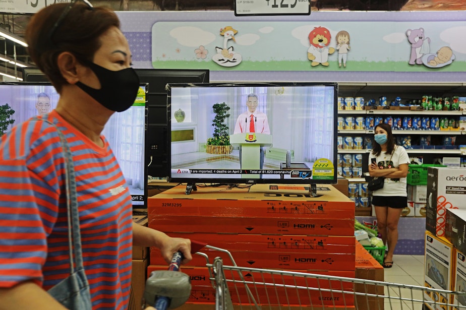 caption: Singapore Prime Minister Lee Hsien Loong addresses the country via live telecast while customers shop for groceries on April 3. Early in the outbreak, Singapore did not recommend masks for the public because of shortages, but began urging public mask-wearing this month to prevent transmission by infected people who were not showing symptoms.