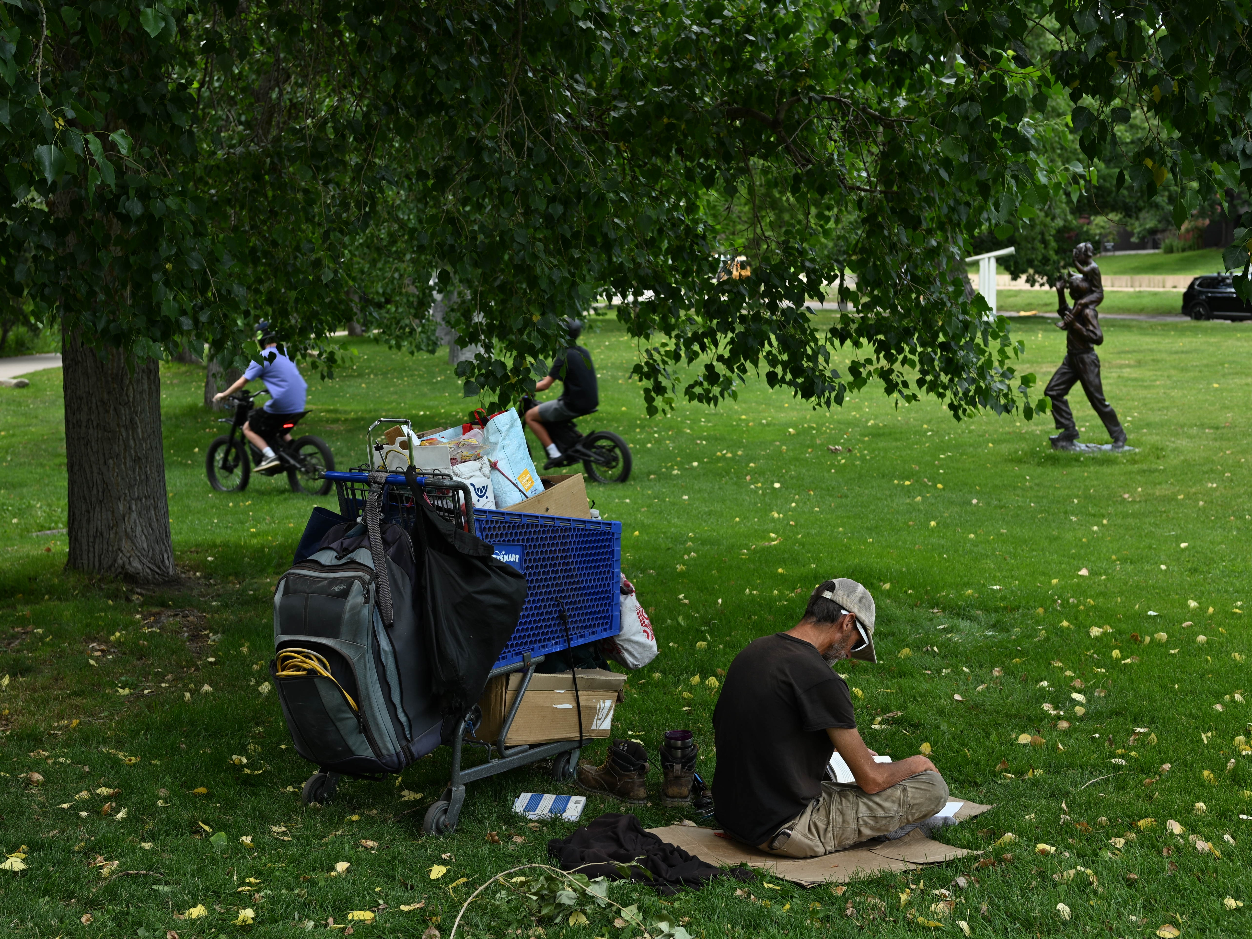 caption: A man who identifies as homeless reads a book under the shade of a tree during high 90-degree temperatures last month in Boulder, Colo.