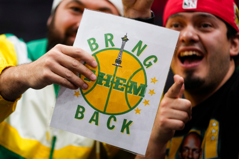 caption: Fans pose for a photo with a sign calling to bring the Seattle SuperSonics back to Seattle, during the first half of an NCAA college basketball game between Gonzaga and UConn, Friday, Dec. 15, 2023, in Seattle. 