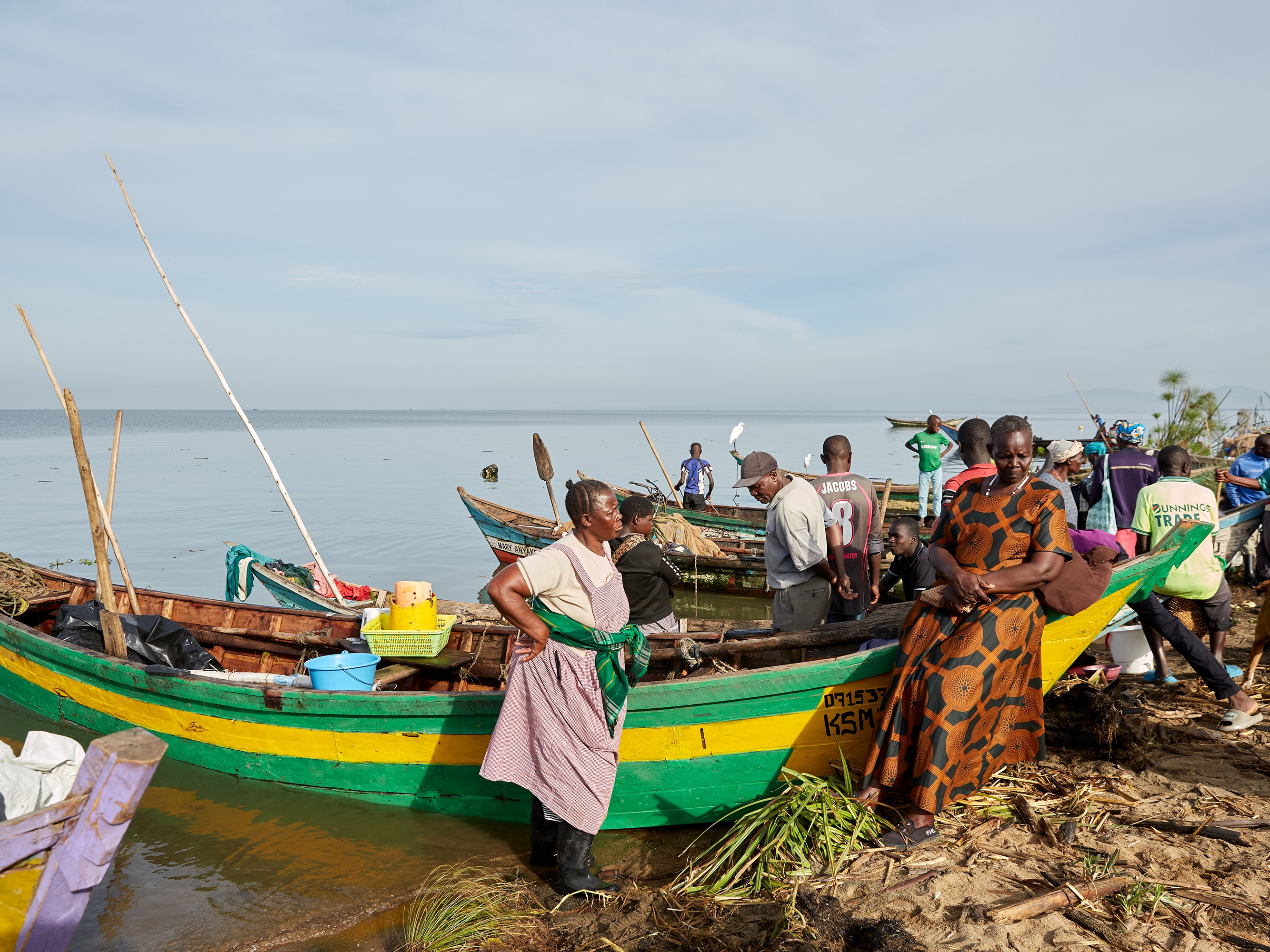 caption: Alice Akinyi (left, hand on hip) and Justine Adhiambo Obura, members of the No Sex For Fish group, stand next to Alice's fishing boat at Nduru Beach. It's one of the few boats that have weathered the storms and floods that have beset the area since 2020.