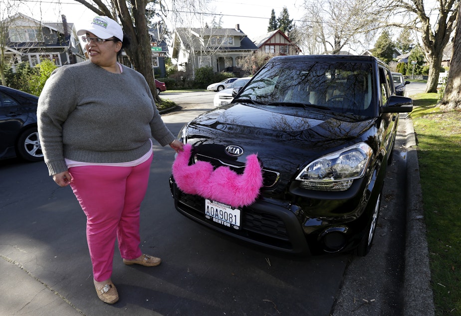 caption: Dara Jenkins, a driver for the ridesharing service Lyft, adjusts the furry mustache drivers display on their cars when they are working, as she starts a shift in Seattle.