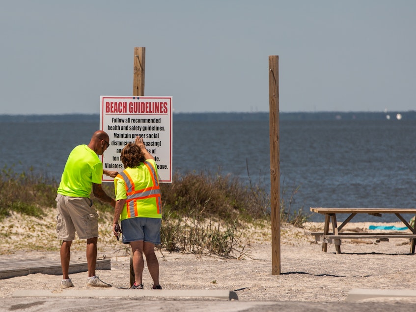 caption: Public workers install a "Beach Guidelines" sign displaying new social distancing rules at a public beach in Dauphin Island, Ala., on Friday. Alabama has reopened some businesses, with restrictions.