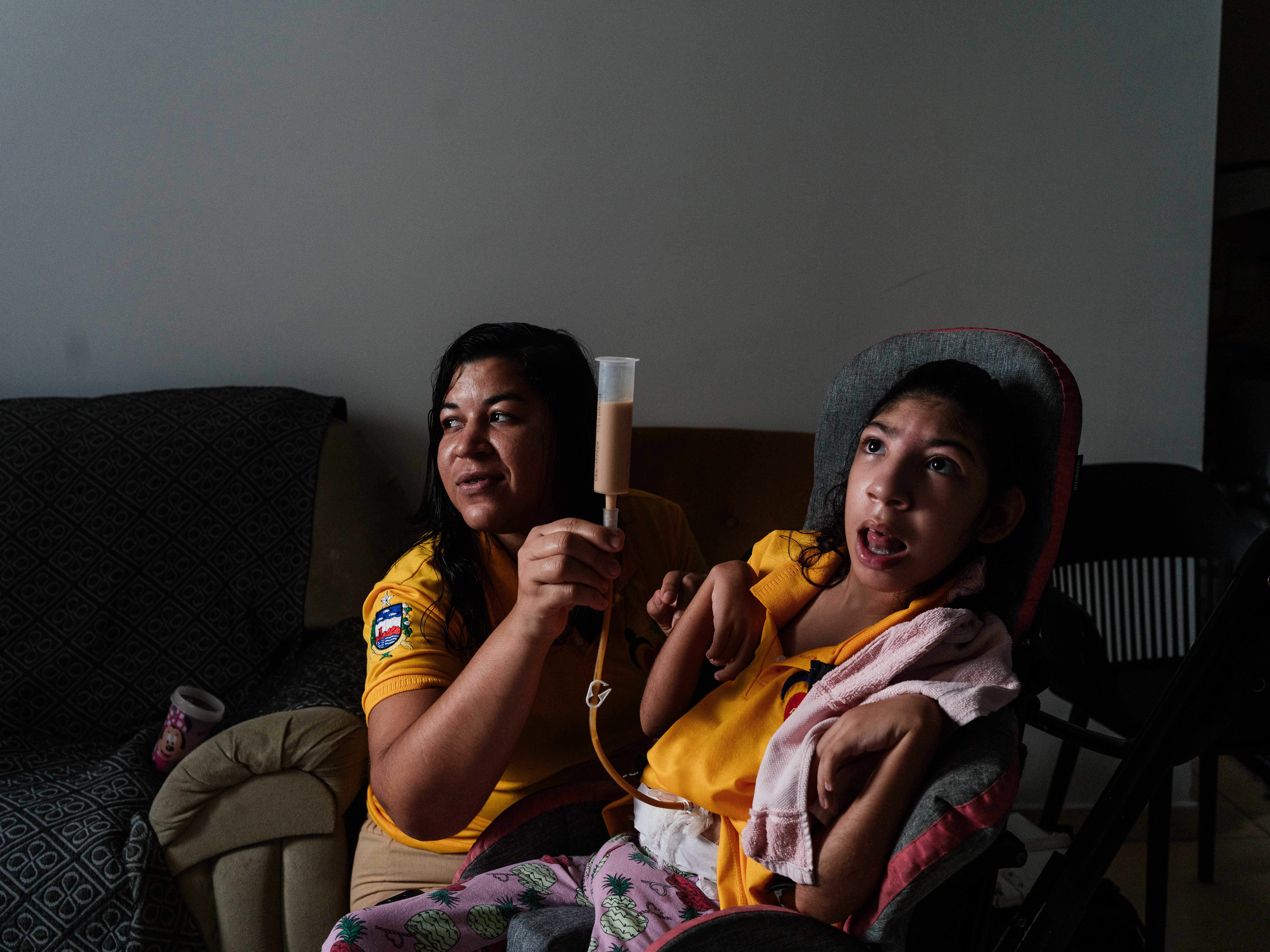 caption: Ruty Pereira sits with her daughter, Tamara, in their apartment in Maceió, in eastern Brazil. Tamara, whose development was impacted in utero by the Zika virus, eats through a feeding tube.