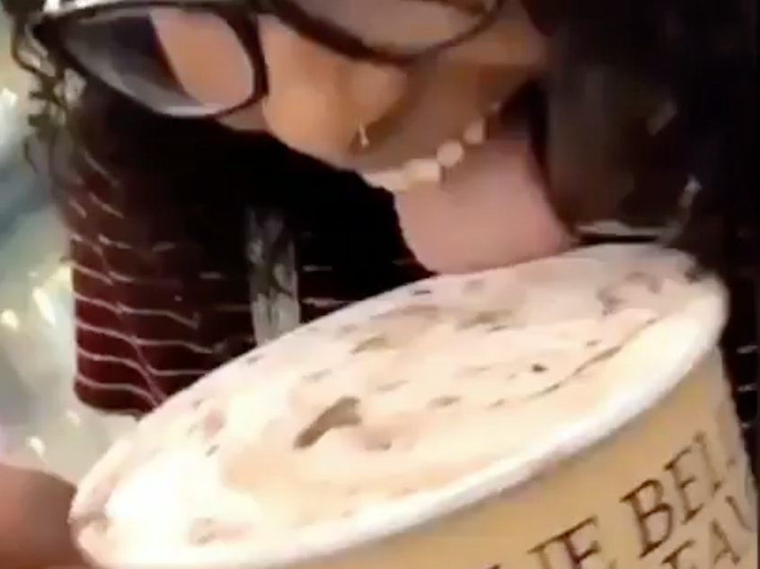 caption: A screenshot of a video shows a young woman opening a carton of ice cream, licking it and putting it back in the grocery store case.