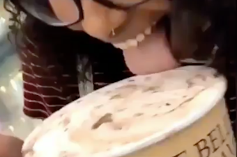 caption: A screenshot of a video shows a young woman opening a carton of ice cream, licking it and putting it back in the grocery store case.