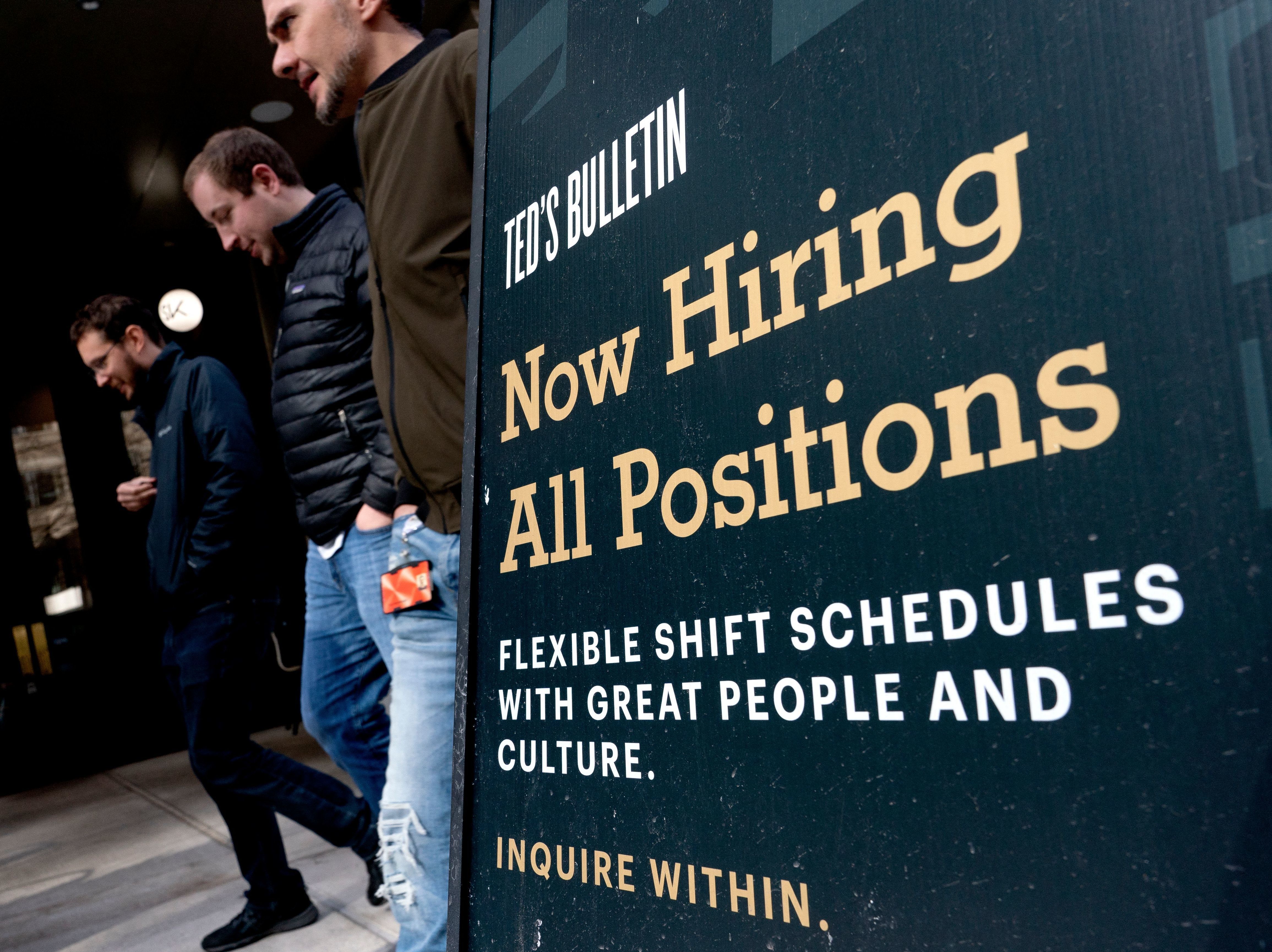 caption: Pedestrians walk past a Now Hiring sign in Arlington, Virginia, on March 16.