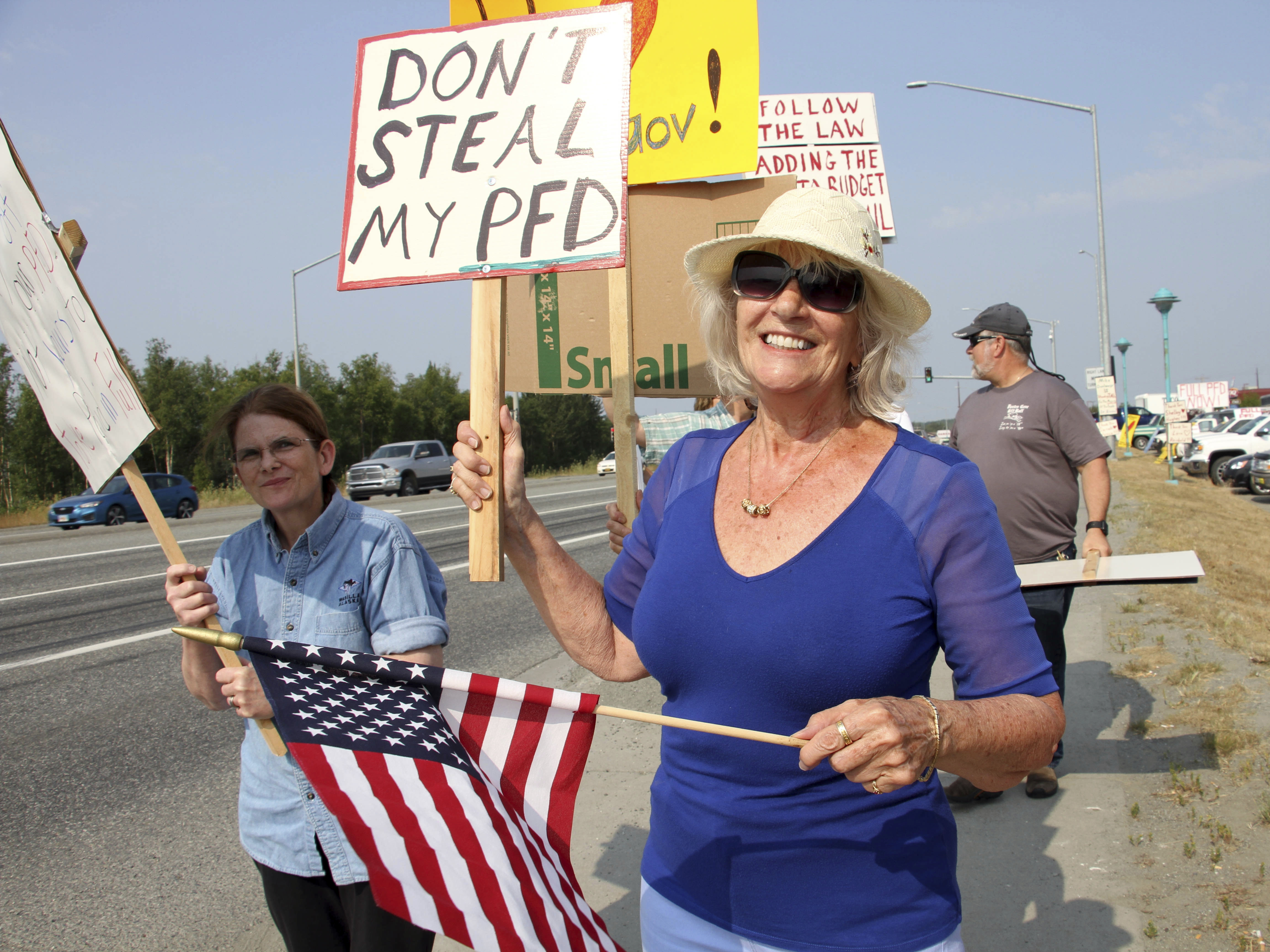 caption: Katherine Hayes waves a flag and a sign in Wasilla, Alaska, urging state lawmakers to fund a full oil wealth fund check, known locally as the PFD for Permanent Fund Dividend.