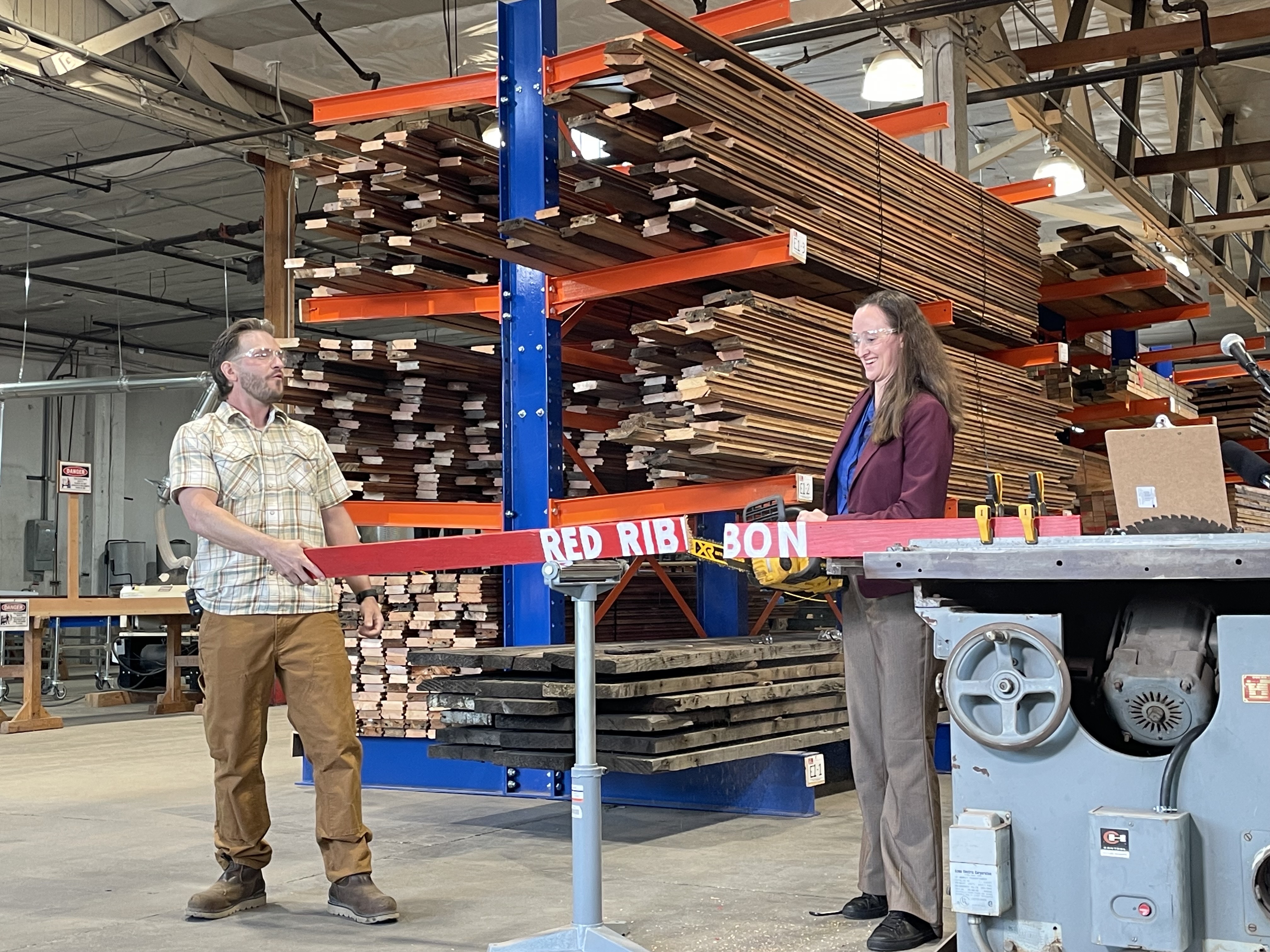 caption: Seattle Mayor Katie Wilson uses a chainsaw to cut a wooden ribbon to celebrate the opening of the city's first lumber salvage warehouse. Aaron Blanchard of Earthwise assists.
