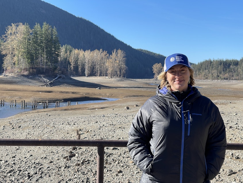 caption: Julie Crittenden of Seattle Public Utilities stands before a desiccated stretch of a reservoir on the Cedar River on Nov. 17, 2023.