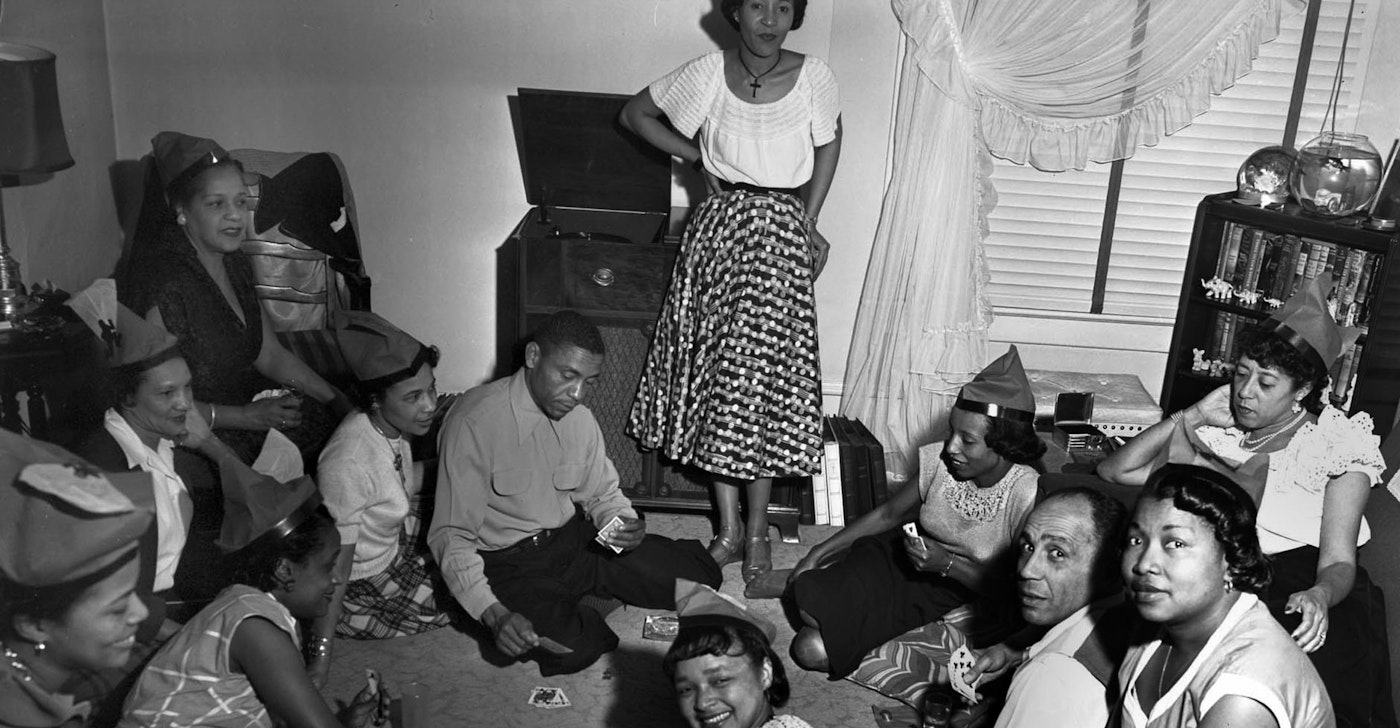 caption: Women, children and two men seated around a living room playing cards. The women wear identical party hats (might this be a birthday?). Identifications: Possibly Glenn Buxton, Cecilia "Babe" Homes, Margie Henderson, Bruce Rowels, Madelaine Brown.
