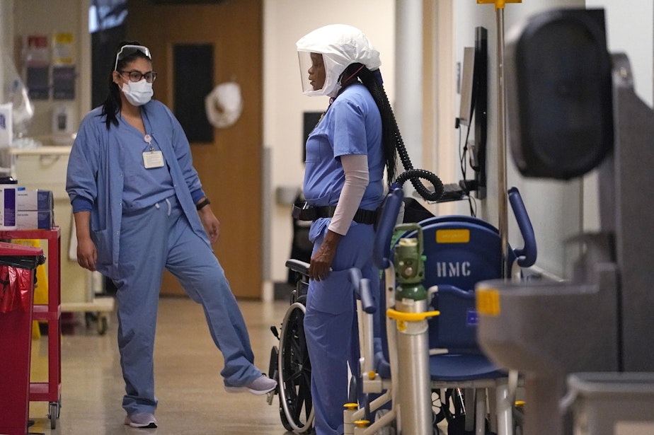 caption: Hospital assistant Tracy Chambers, right, talks with another staff member in a hallway in the acute care unit, where about half the patients are COVID-19 positive or in quarantine after exposure, of Harborview Medical Center, Friday, Jan. 14, 2022, in Seattle. 