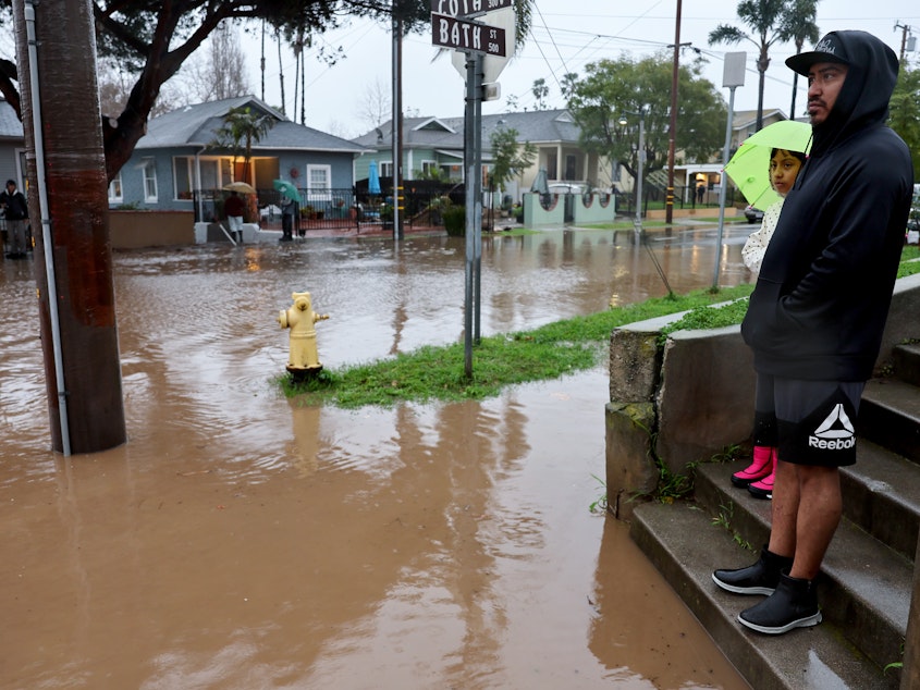 caption: Residents stand along a flooded street in Santa Barbara, California, as a powerful atmospheric river pummels the region. The storm has caused landslides, power outages, and road and airport closures across Southern California.