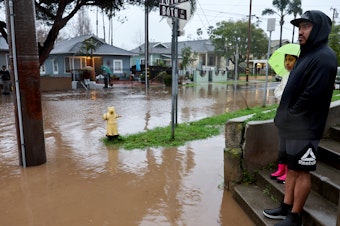 caption: Residents stand along a flooded street in Santa Barbara, California, as a powerful atmospheric river pummels the region. The storm has caused landslides, power outages, and road and airport closures across Southern California.