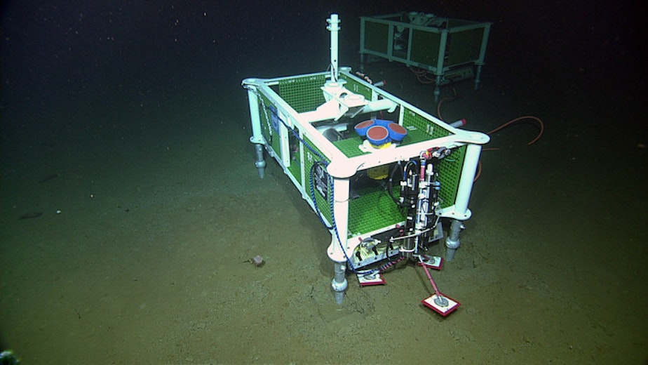 caption: An Acousitic Doppler Current Profiler (four blue-red cyllinders with yellow housing) is installed at the base of Axial Seamount.