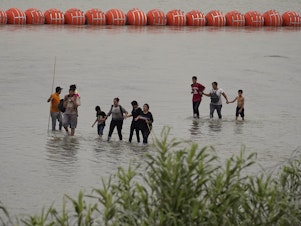 caption: Migrants who crossed the Rio Grande from Mexico walk past large buoys being deployed as a border barrier on the river in Eagle Pass, Texas, on Wednesday.