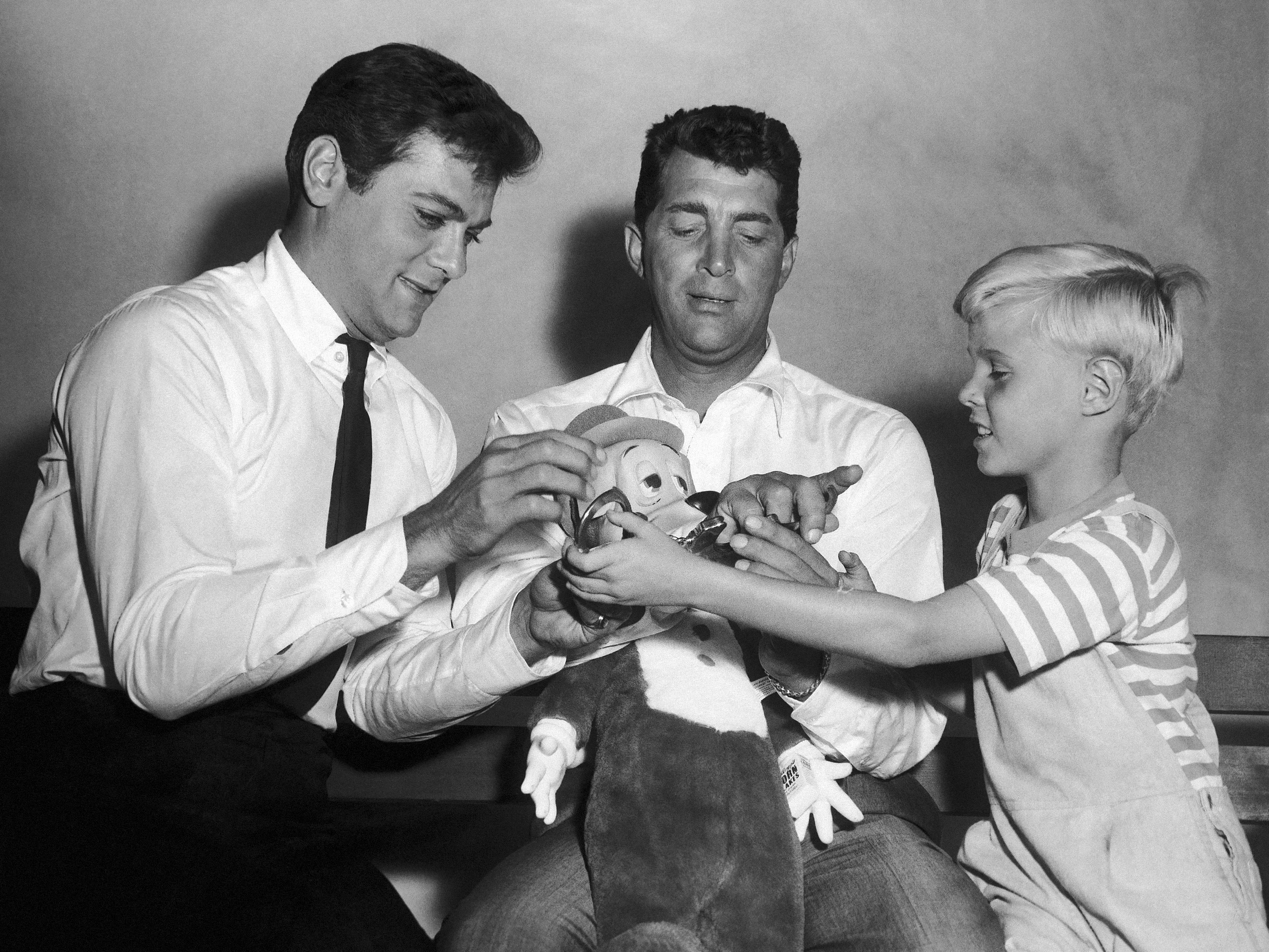 caption: Jay North, young star of the new <em>Dennis the Menace</em> series, right, attempts to handcuff Dean Martin, center, as they play with a stuffed toy with Tony Curtis on Sept. 15, 1959.