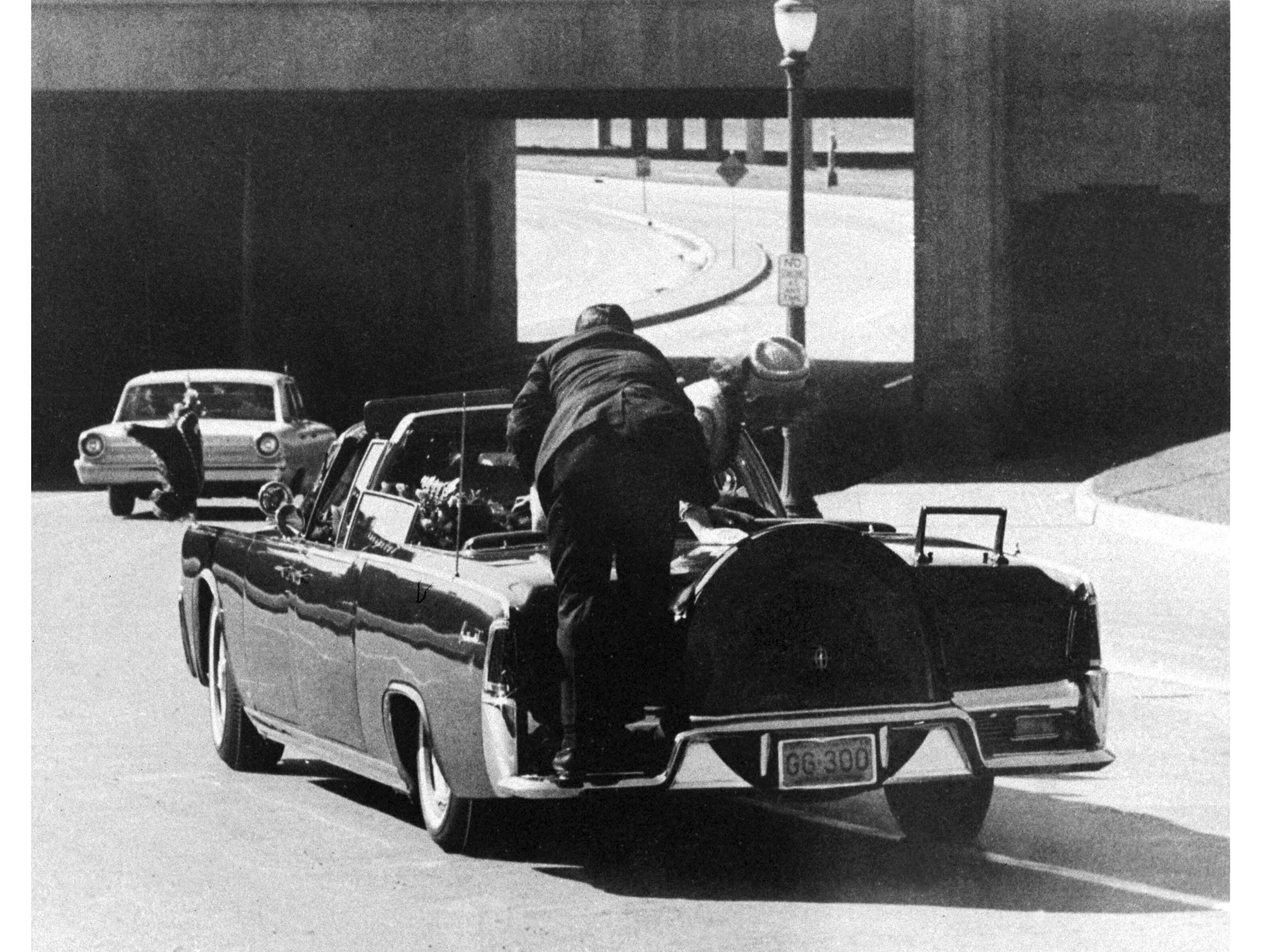 caption: President John F. Kennedy slumps down in the back seat of the presidential limousine as it speeds along Elm Street toward the Stemmons Freeway overpass in Dallas, Texas, after being fatally shot, Nov. 22, 1963. First lady Jacqueline Kennedy leans over the president as Secret Service Agent Clint Hill pushes her back to her seat.