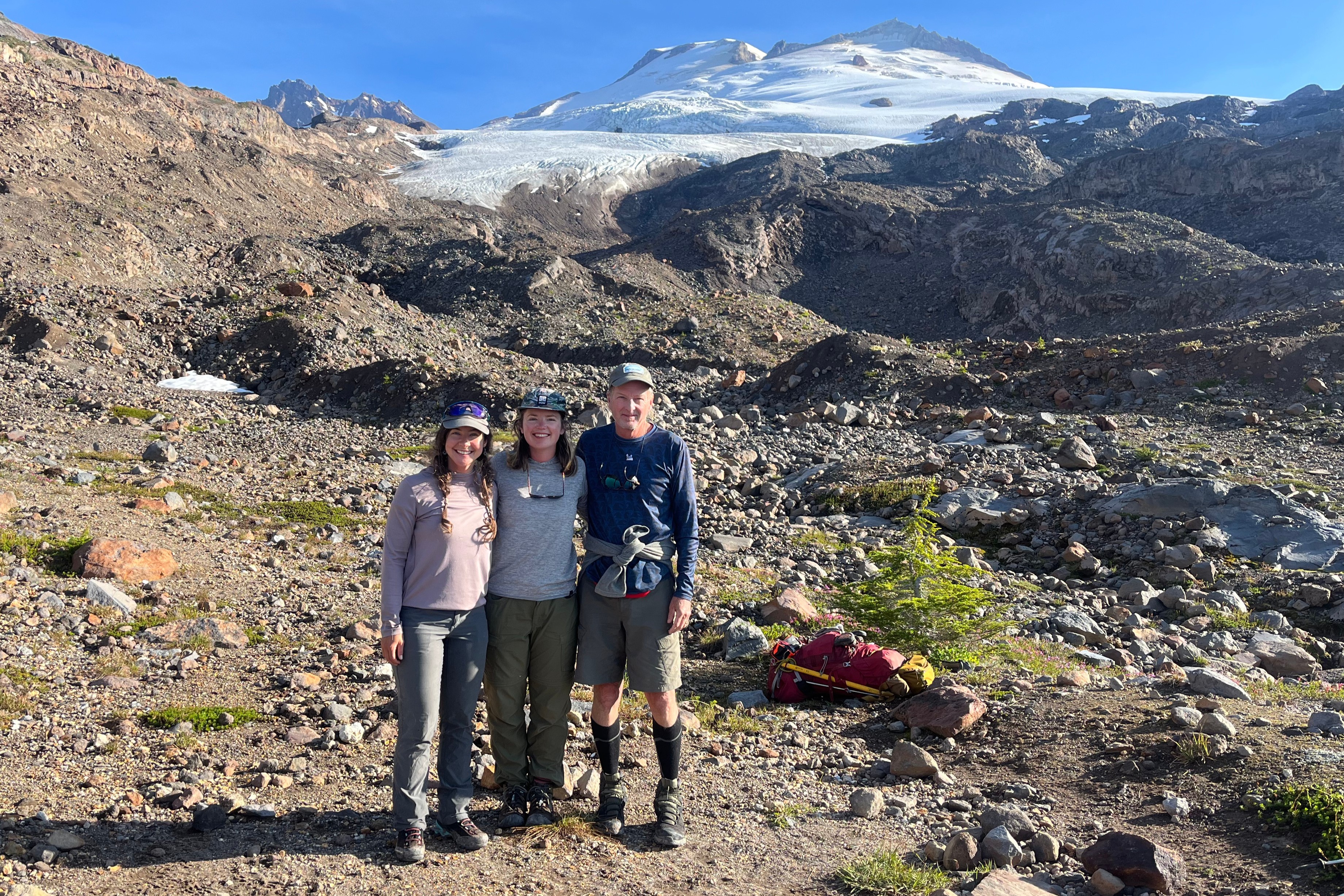 caption: Glacier researchers Abby Hudak, Jill Pelto and Mauri Pelto pose beneath a dwindled Easton Glacier on Washington's Mount Baker on Aug. 9, 2025.