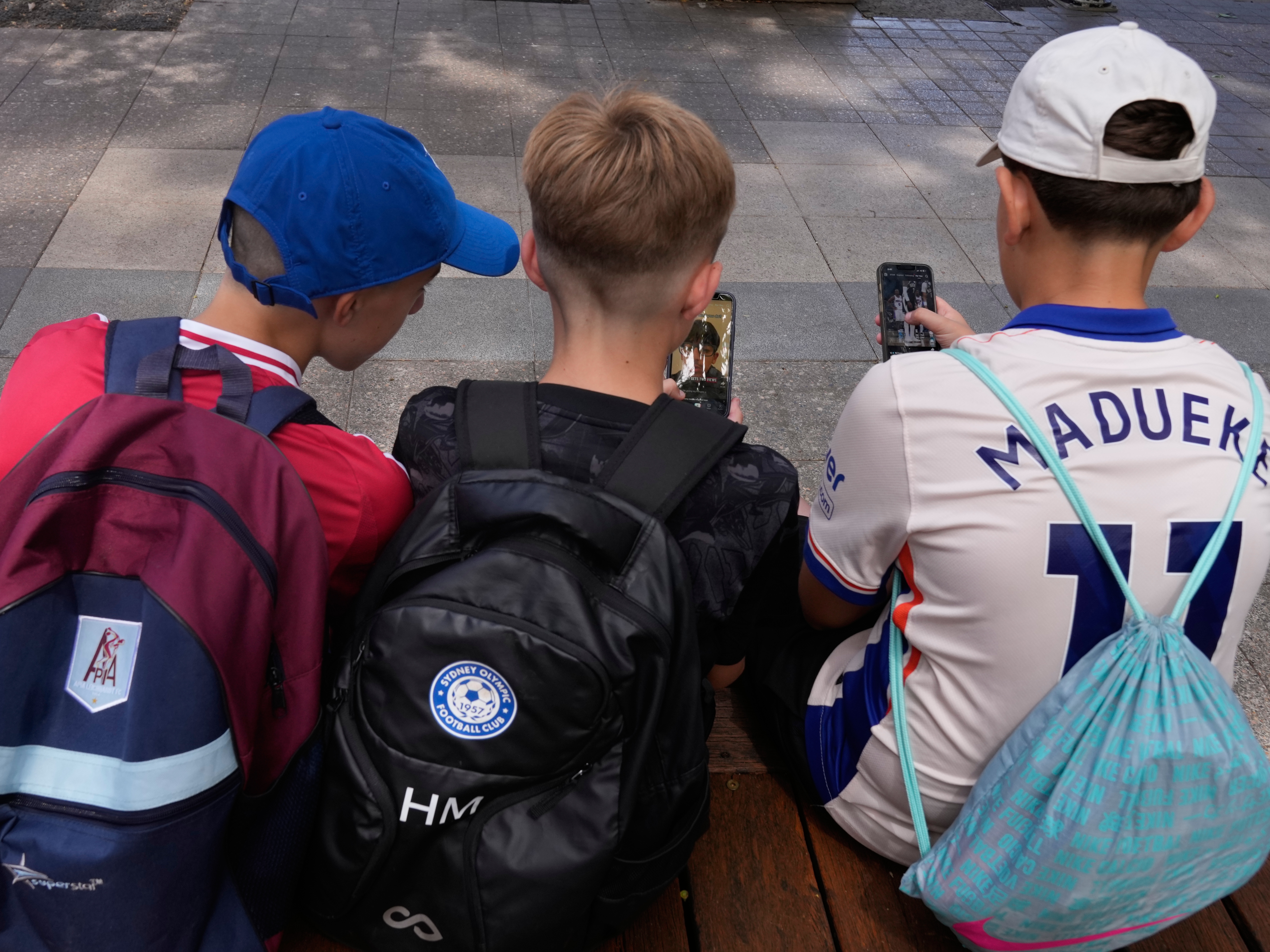 caption: Hugo Winwood-Smith, right, Hardy Macpherson and Edan Abou, left, all 11-years-old, use their phones while sitting outside a school in Sydney on Monday, Dec. 8, 2025.